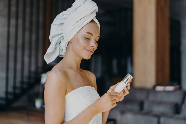 Pleased healthy woman with smooth skin, minimal makeup, holds bottle of body lotion, wrapped in bath towel, poses indoor against blurred background. Female with skin care product. Beauty concept - Foto, Imagen