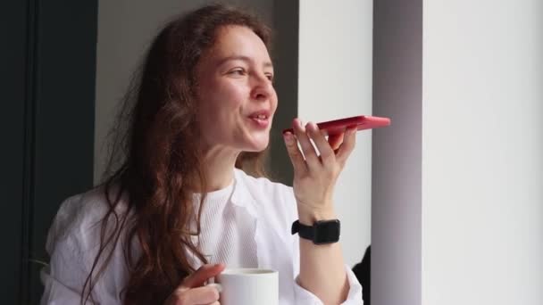 Beautiful millennial woman recording answering by audio message in social network for friend, sending voicemail using virtual app on mobile phone, standing near window with cup of coffee at home. - Imágenes, Vídeo