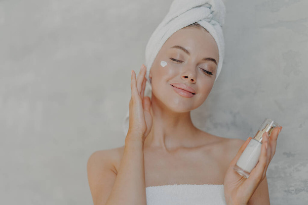 Headshot of pleased attractive woman applies face lotion, satisfied with new cosmetic product, keeps eyes closed, touches soft skin after bath, has well cared complexion, poses against grey wall - Photo, image
