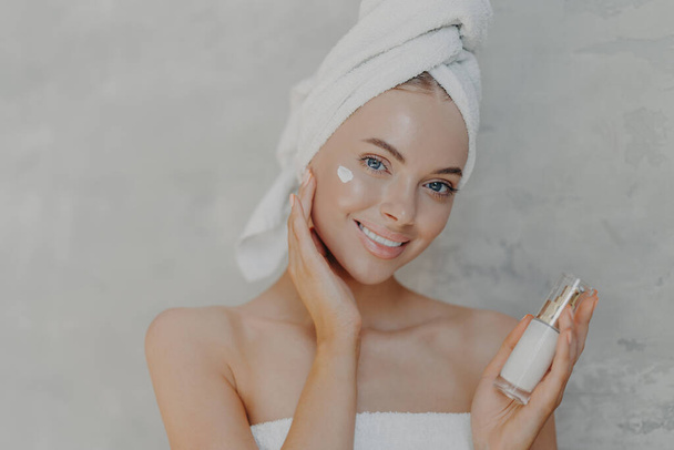 Close up shot of pretty young European woman with minimal makeup, applies moisturising cream on face, smiles gently and looks at camera, wears bath towel on head, isolated over grey background - Photo, Image