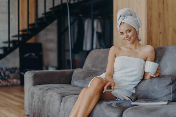 Smiling relaxed young European woman sits on comfortable sofa, reads magazine and drinks coffee, poses on comfortable sofa, undergoes beauty treatments, has satisfied expression. Spending time at home - Φωτογραφία, εικόνα
