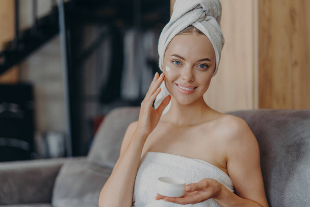 Pretty young woman with healthy smooth skin applies face cream, wears wrapped towel on head after taking shower, poses on comfortable sofa. Hygiene procedure, cosmetology and beauty concept. - Photo, Image