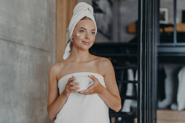 Thoughtful lovely young female model with cream on face, wrapped in towel, concentrated somewhere aside, holds mug of tea, undergoes beauty treatments at home, stands indoor. Body care concept - Φωτογραφία, εικόνα
