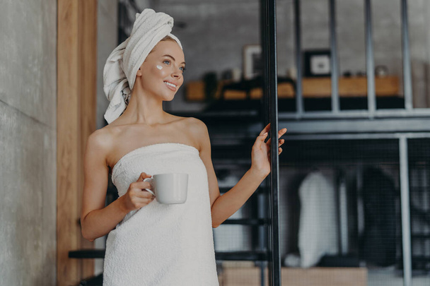 Pretty thoughtful woman concentrated somewhere, smiles broadly, has white perfect teeth, applies face cream, stands wrapped in towel, drinks hot tea, stands on stairs indoor, relaxes after taking bath - Zdjęcie, obraz
