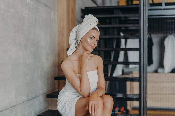 Thoughtful young European woman keeps hand under chin, sits on stairs and has thoughtful expression, has healthy skin, puts face cream, wrapped in bath towel, undergoes beauty treatments at home - Zdjęcie, obraz