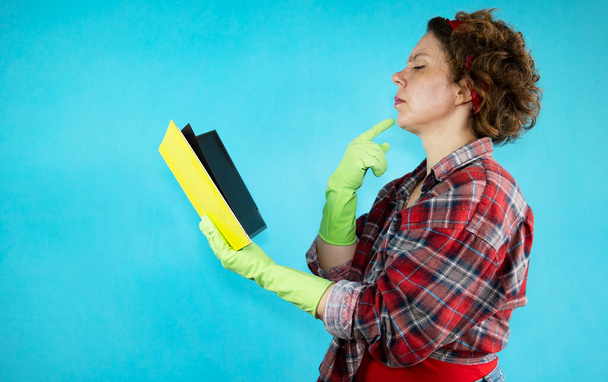 Pensive pin-up woman looking at yellow book on isolated blue background. The woman is reading. Woman cleaning in green gloves - Foto, afbeelding