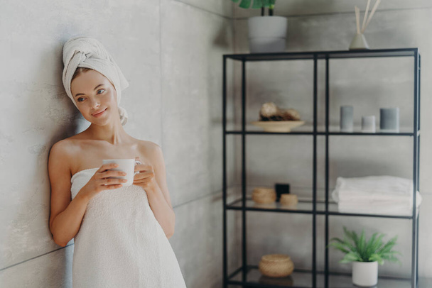 Kuva rauhallinen rento nainen aiheuttaa pyyhkeessä vastaan kylpyhuoneen sisustus, kääritty kylpypyyhe, juo teetä tai kahvia, on puhdas täydellinen iho, nojaa harmaa seinä. Kauneuspäivä, kylpylä ja ihonhoito - Valokuva, kuva