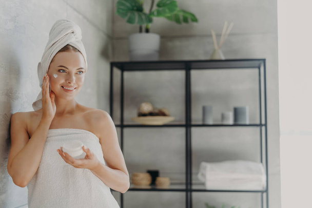 Lovely smiling Caucasian woman applies beauty cream on cheek, enjoys morning domestic skin care routine, wrapped in bath towel, grooming herself after showering poses in bathroom. Hygiene concept - Photo, Image