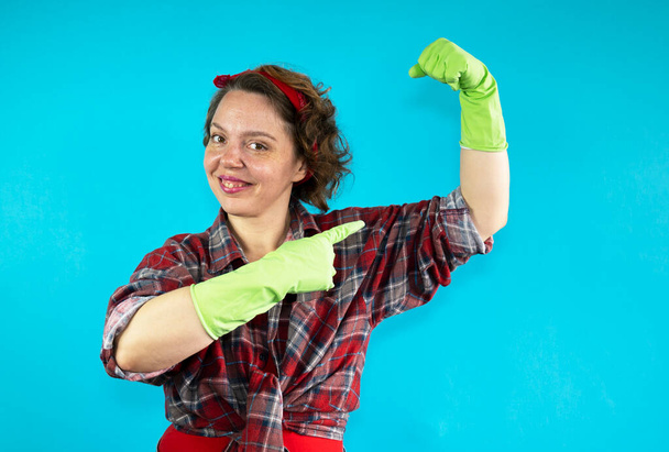 Μια ενήλικη γυναίκα με καρό πουκάμισο δείχνει ένα δάχτυλο στους δικέφαλους με πράσινα λαστιχένια γάντια σε ένα μπλε απομονωμένο φόντο. Καθαρισμός γυναικών. Καρφιτσώστε στυλ. Γυναικείο πορτρέτο - Φωτογραφία, εικόνα