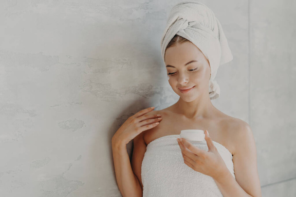 Beautiful satisfied woman with healthy skin, minimal makeup, applies face cream, stands wrapped in bath towel near grey background after taking shower. Sauna morning routine, skin care concept - Photo, Image