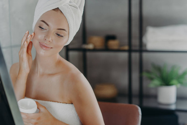 Υπέροχη υγιής γυναίκα εφαρμόζει ενυδατική κρέμα, τυλιγμένο σε πετσέτα μπάνιου, κοιτάζει προσεκτικά την αντανάκλαση στον καθρέφτη, απολαμβάνει τη θεραπεία φροντίδας του δέρματος μετά το ντους, θέτει στο μπάνιο. Έννοια ομορφιάς. - Φωτογραφία, εικόνα
