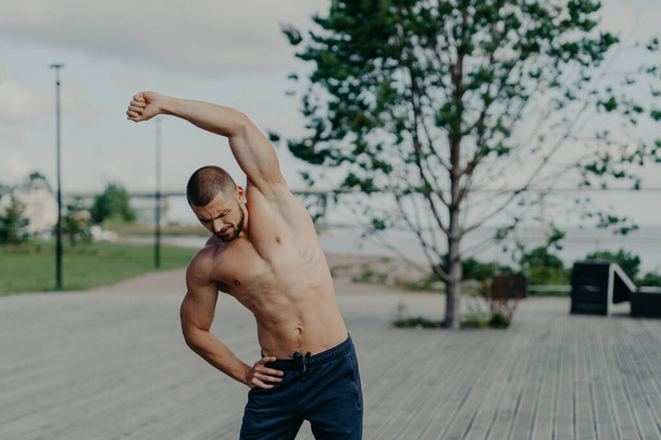 Active sportsman with naked torso and muscular body, does stretching exercises outdoor, shows good flexibility. Motivated athlete unshaven man warms up, prepares for workout, keeps muscles flexible. - Foto, Bild