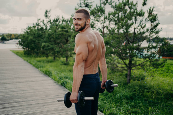 Muscular satisfied athlete bearded man gets energy during sport training, has strong arms, raises barbells, dressed in shorts, poses outdoor, enjoys regular workout in nature, builds healthy body. - Photo, Image