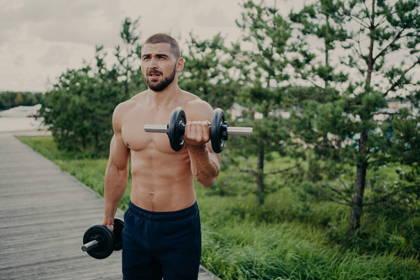Body care and power lifting concept. Strong muscular athlete bearded man raises dumbbells, lifts heavy barbells, performs shoulder press, poses outside against nature background. Sport exercising - Photo, Image