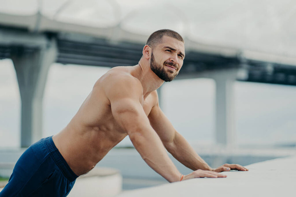Boční pohled na atletický motivovaný cílevědomý vousatý sportovní nahý hrudník muž provádí push-up cvičení, dělá úsilí opřený o most, proto se dívá do dálky, má svalnaté tělo, cvičí venku. - Fotografie, Obrázek
