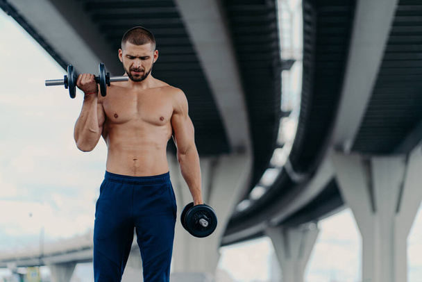 Outdoor shot of strong muscular man raises barbells and trains muscles stands under bridge has aim to have healthy body shape, has fitness training or physical workout. Athletic bodybuilder. - Foto, immagini