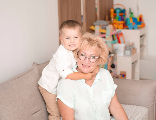 Menschen. Eine Familie. Glückliche und schöne kaukasische Großmutter und kleiner Enkel in weißen Hemden umarmen sich, während sie auf dem Sofa im heimischen Interieur sitzen. Familienkonzept. Weicher Fokus. - Foto, Bild
