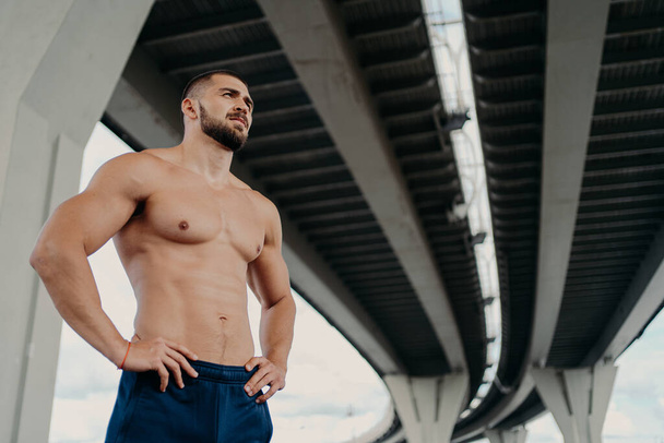 Strong man with naked torso keeps hands on waist, has muscular body after long training, focused into distance, stands under bridge, thinks about how to be healthy and fit. Sporty man outdoor - Photo, Image