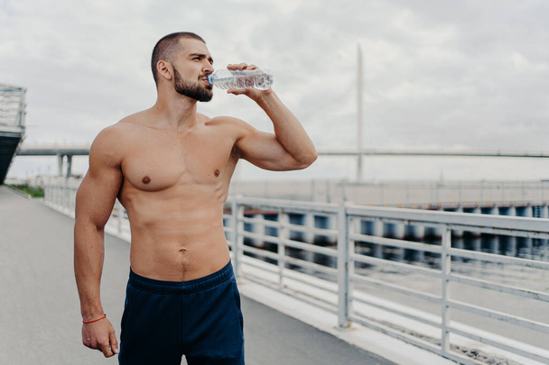 Actieve sportman drinkt fles water poseert met naakte romp geconcentreerd in afstand voelt dorst na het lopen buiten vol energie, leidt een actieve levensstijl. Sport voor een beter leven - Foto, afbeelding