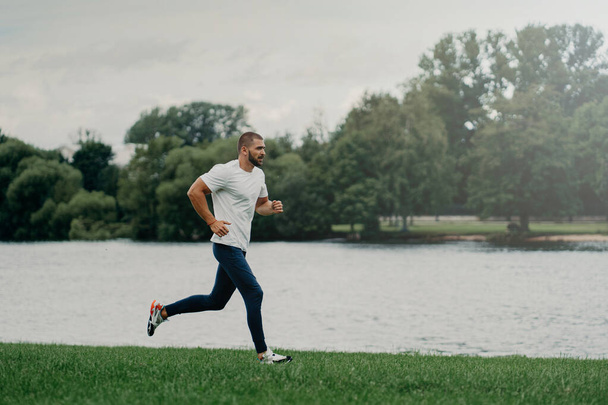 Sporty European man dressed in t shirt, trousers and sneakers and runs along river, trains outdoors, leads active lifestyle, breathes fresh air. People, sport, wellness and recreation concept. - Foto, imagen