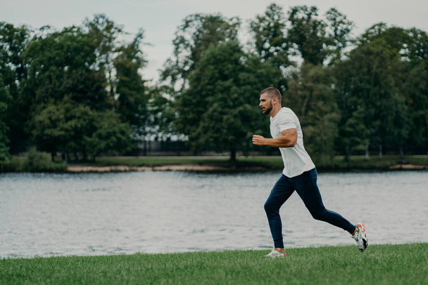 Przystojny sportowiec brodaty mężczyzna biegnie na zewnątrz w godzinach porannych pozy w pobliżu rzeki cieszy się przyrodą i świeżym powietrzem, wykazuje wytrzymałość i motywację, ma poranny trening na co dzień, pozostaje zdrowy silny - Zdjęcie, obraz