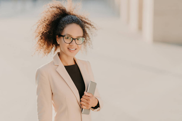 Θετική πανέμορφη Afro American επιχειρηματίας βόλτες εξωτερική με touchpad, φοράει επίσημα ρούχα, οπτικά γυαλιά, χαμόγελα στην κάμερα, απολαμβάνει ελεύθερο χρόνο μετά τη συνάντηση. Η έννοια των ανθρώπων και της τεχνολογίας - Φωτογραφία, εικόνα