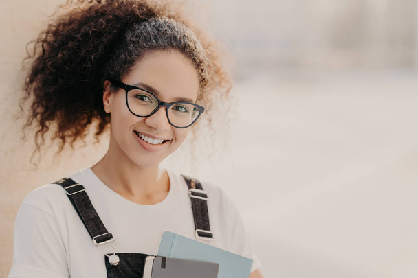 Horizontal shot of beautiful woman with crisp hair combed in pony tail, carries notepad, wears white t shirt and overalls, enjoys studying, poses against blurred background with empty space for text - Foto, afbeelding