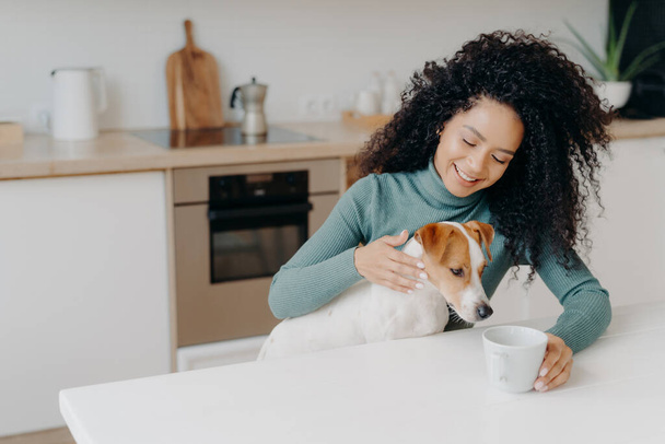 Happy Afro African woman with curly hairstyle treats dog in kitchen, pose at white table with mug of drink, enjoy domestic atmosphere, have breakfast together. People, animals, home concept. - Photo, Image
