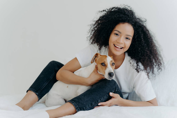 Horizontal shot of glad Afro woman rests in bed with dog, have playful mood, pose together in bedroom against white background. Girl relaxes at home with jack russell terrier. Sweet funny moment - Photo, Image
