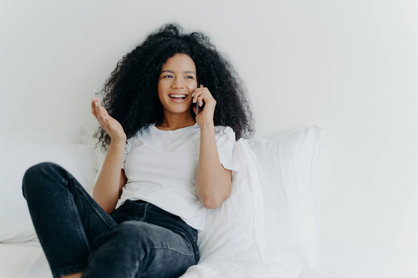 People, technology, communication and rest concept. Glad curly haired ethnic lady has telephone talk, gestures and laughs with joy, looks somewhere aside, spends time at home, sits on white bed - Foto, imagen