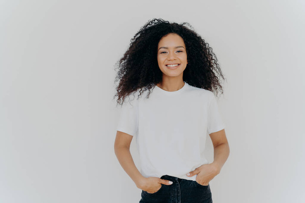 Ізольований знімок молодої афроамериканської жінки носить білу футболку, виражає добрі емоції, стоїть на самоті в приміщенні, подає фотографії, має випадкову розмову з другом, насолоджується вільним часом. Люди і щастя - Фото, зображення