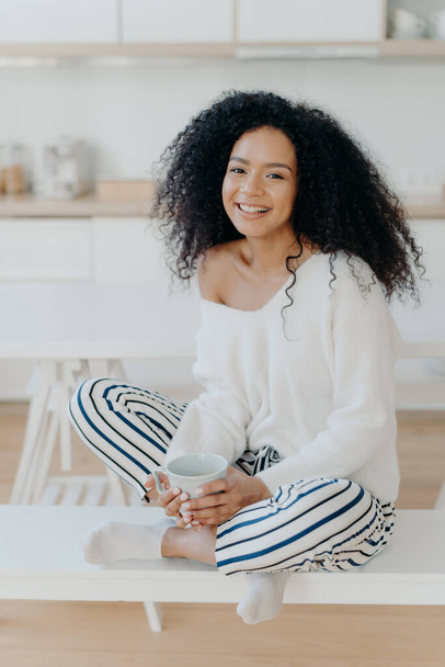 Image of lovely curly haired lady drinks coffee or tea from white mug, wears fashionable white sweater, striped pants, poses at kitchen against blurred background. People and lifestyle concept - Foto, afbeelding