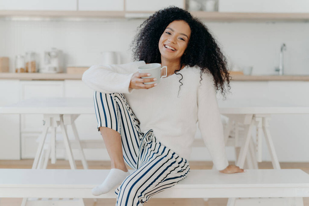 Beautiful young female with Afro haircut, poses on white bench indoor, dressed in stylish clothing, enjoys aromatic beverage, enjoys breakfast at home, poses against blurred kitchen interior - Φωτογραφία, εικόνα