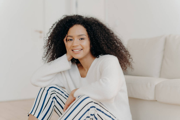 Photo of relaxed delighted African American woman with curly hairstyle, smiles gently at camera, wears white sweater and striped trousers, sits on floor near couch in living room of modern apartment - Фото, зображення