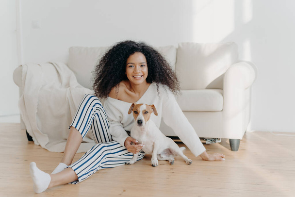 Relaxed young African American woman sits on floor in empty room near sofa with pedigree dog, enjoys sweet moment at home, move in new apartment to live. Lady rests with puppy in living room - Foto, Imagen