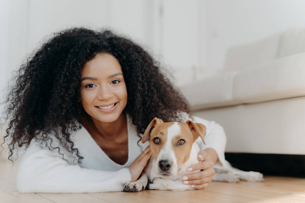 Pretty girl with Afro hair, lies on floor with dog, expresses pleasant emotions, poses in living room near couch, bought pet in new apartment. Woman host with beloved animal at home, share good moment - Foto, afbeelding