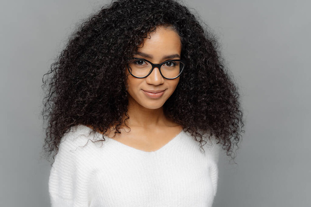 Close up shot of dark skinned female has satisfied expression, bushy curly hair, wears spectacles and white jumper, looks straightly at camera, models over grey background. Natural beauty concept - Photo, Image