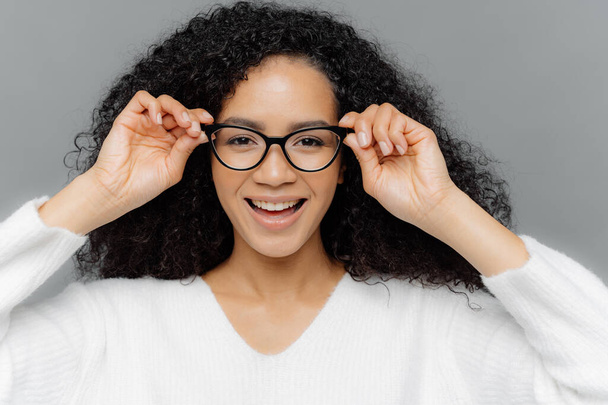 Close up shot of pretty dark skinned female has minimal makeup, keeps hands on frame of transparent glasses, dressed casually, enjoys spare time and good day, isolated over grey studio wall. - Foto, imagen