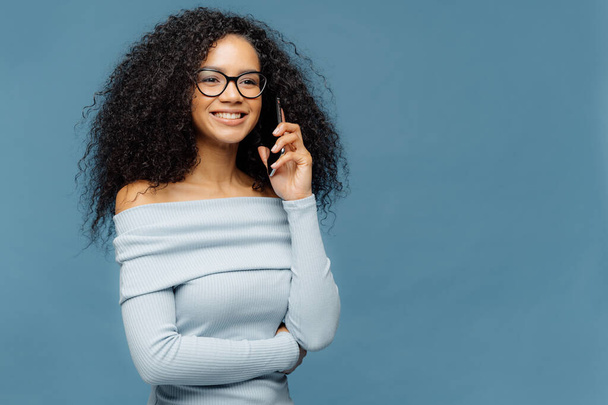 Smiling delighted Afro American woman with curly hair, talks on smart phone, discusses some pleasant moments after party, looks happily into distance, wears glasses and jumper, isolated on blue wall - Fotoğraf, Görsel