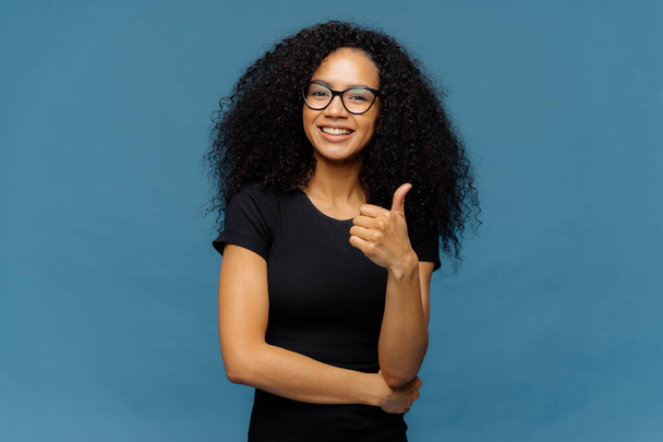 Photo of lovely dark skinned female makes like gesture, keeps thumb up, approves something good, has pleasant smile, wears spectacles and casual black t shirt, isolated over blue background. - Photo, Image