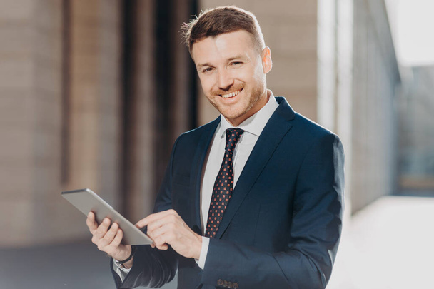 Photo of successful male banker does remote work on digital tablet, has happy expression, dressed in formal black suit, has happy look at camera. Positive young man makes transactions online - Photo, Image
