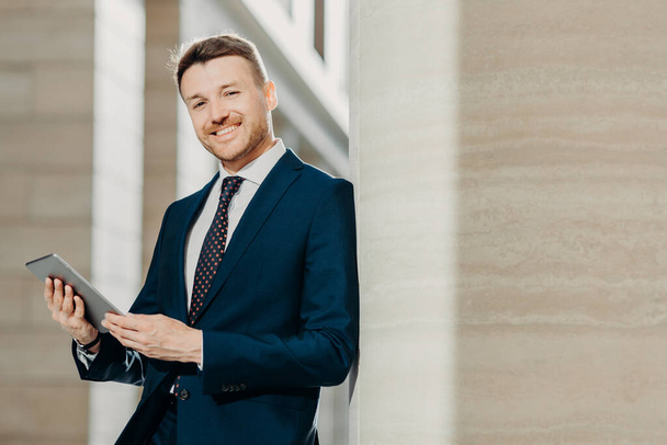 Portrait of stylish male entrepreneur has cheerful expression, holds digital tablet, reads financial news on web page, uses free internet connection, wears formal suit and tie. Career concept - Photo, Image