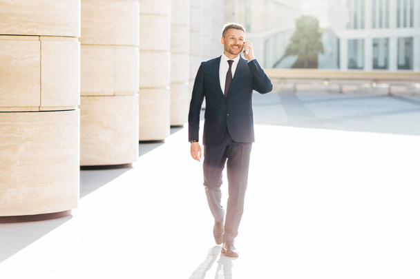 Prosperous male business owner in formal suit, has telephone conversation with business partner while walks to meeting in urban setting, satisfied to hear good news. People and career concept - Photo, image