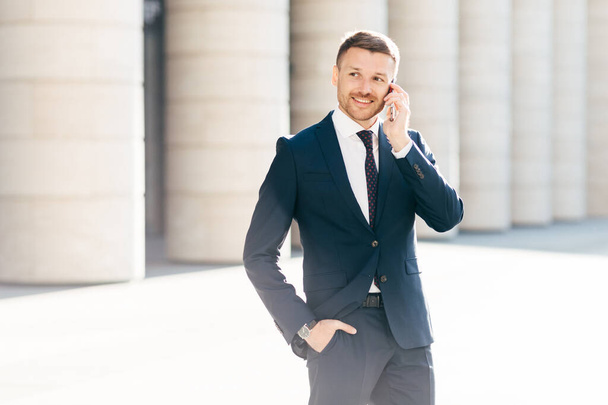 Portrait of successful male manager satisfied with mobile tariffs, makes phone call, uses banking service operator, wears formal suit, stands in urban setting outsie. Professional man worker - Fotoğraf, Görsel