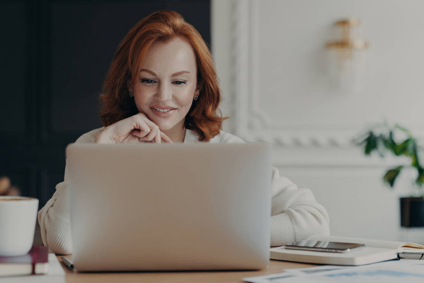 Experienced red haired businesswoman reads publication on modern laptop computer, concentrated at screen with pleasant smile, makes research for planning startup, checks mail box, works from home - Photo, Image