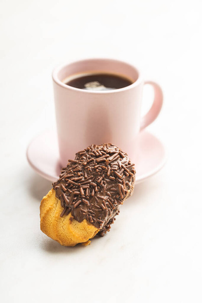 Petit fours with chocolate sprinkles. Mini chocolate dessert and a coffee cup on white kitchen table. - Фото, изображение