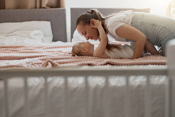 Молодая мама улыбается и целует своего ребенка, лежа дома в постели. Размытый фон - Фото, изображение