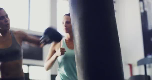 Female athlete boxer, exercising with a punching bag with coach watching at training fitness gym. Active sport woman with boxing gloves doing workout fighting routine with instructor indoors - Imágenes, Vídeo