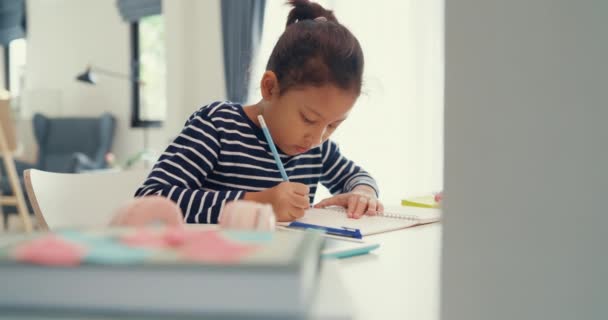 Азиатская малышка в свитере сидит перед столом с блокнотом с помощью карандаша сосредоточиться на записи ноутбука делать домашнее задание из онлайн-курса обучения в выходные дни дома. Дистанционное онлайн обучение. - Кадры, видео
