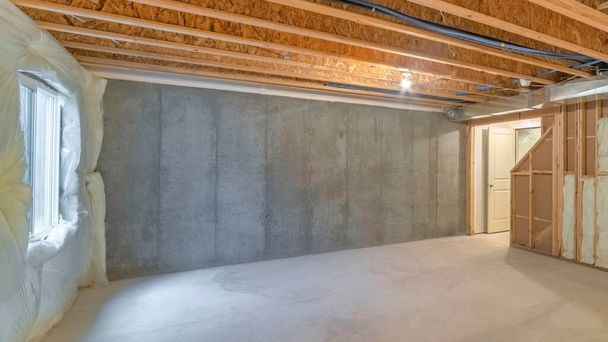 Panorama Unfinished basement with a plastic vapor barrier on the wall. Empty basement interior with wooden frameworks and insulated wall on the left and a window on the right. - Photo, Image